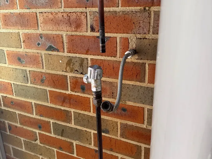 Water pipes, a braided hose and an isolation valve disconnected on a brick wall beside a hot water system in Harrington Park