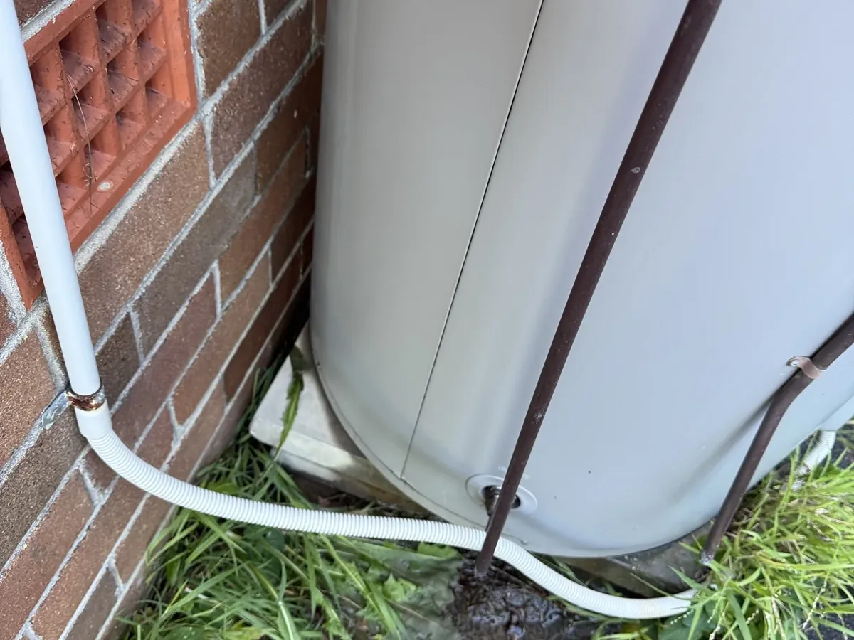 Wet ground and a drain line at the base of a leaking storage tank beside a brick wall in Riverwood