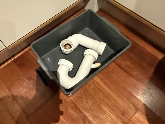 Sink trap taken apart and set in a grey tray on a timber floor beside kitchen cupboards in Erskineville