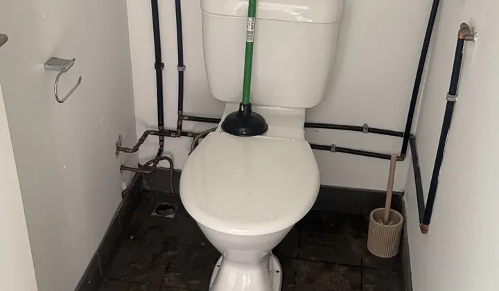Plunger resting on the lid of a warehouse toilet with exposed water pipes in Minto