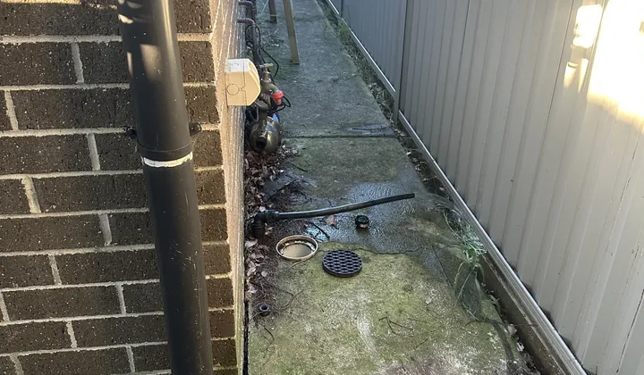 Drain opening and a lifted grate on a mossy side path beside a brick wall in Mount Druitt