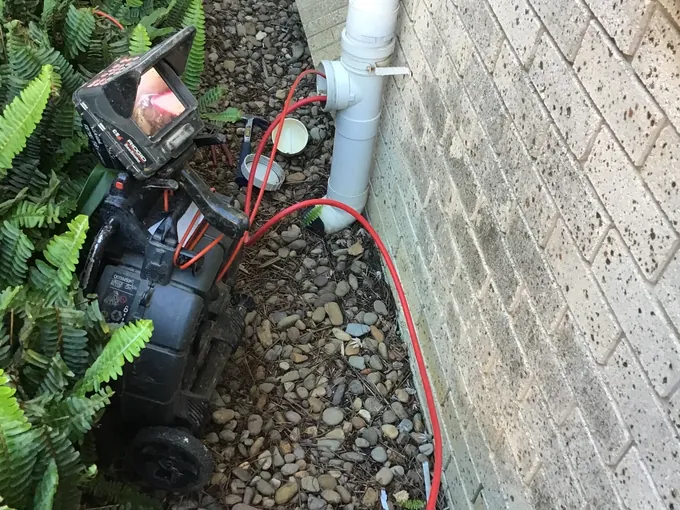 Red jetter hose run into the inspection opening on a white drain pipe, with a drain camera beside it, in Baulkham Hills