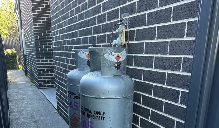Two tall LPG gas bottles and their regulator on a dark brick wall beside a side path in Condell Park