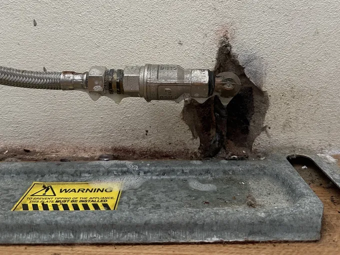 Braided gas hose and valve at the wall behind a freestanding stove, above its anti-tip plate, in Collaroy