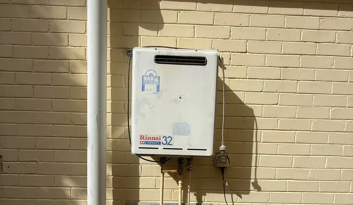 Weathered continuous flow gas hot water unit mounted on a cream brick wall between downpipes in Oatley