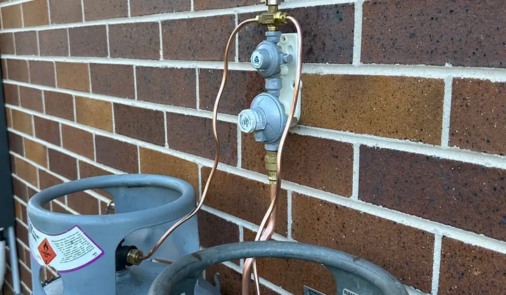 Two LPG gas bottles against a brick wall with new copper pigtails running to a changeover regulator, in Berowra Heights