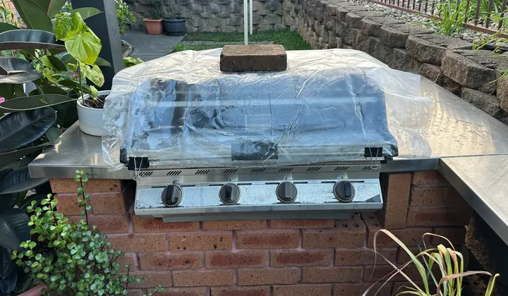 Built-in BBQ under a plastic cover in a brick outdoor kitchen with a garden behind in Quakers Hill
