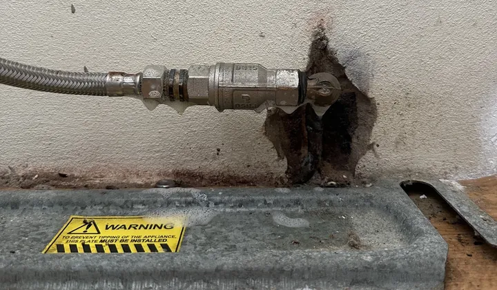 Braided gas hose and valve at the wall behind a freestanding stove, above its anti-tip plate, in Collaroy