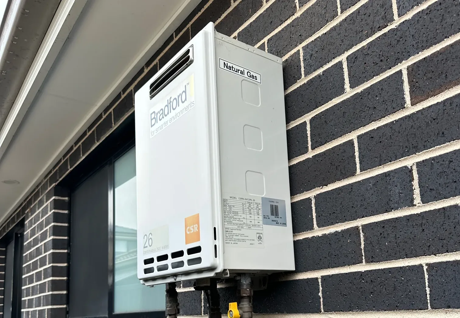 Bradford continuous flow gas hot water unit with copper pipes and a gas valve on a black brick wall in Austral