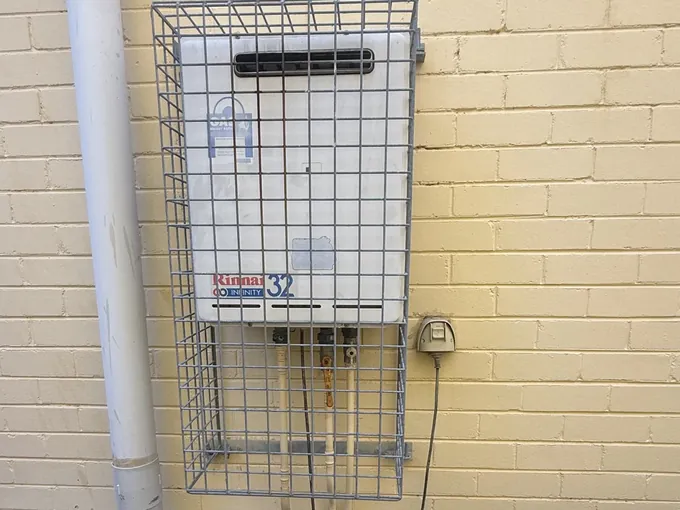 Continuous flow gas hot water unit in a wire cage on a brick wall in Oatley