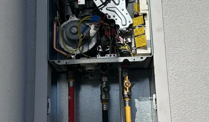 Gas continuous flow unit with its cover off in a recess box above red, black and yellow pipes in Spring Farm
