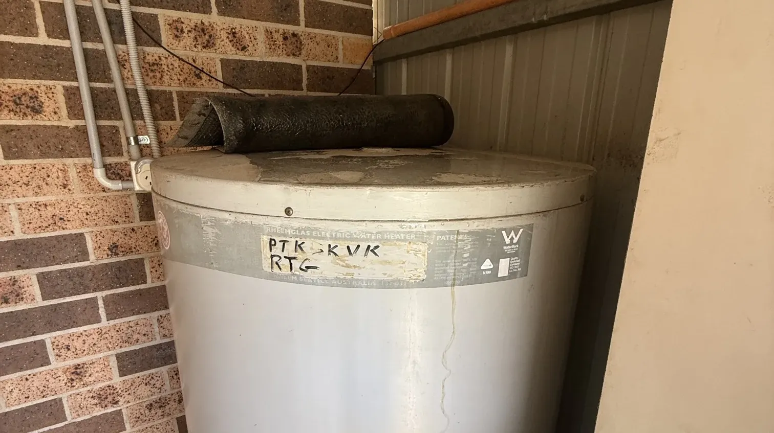 Old electric hot water tank with a water stain running down its side under a lean-to roof in Austral