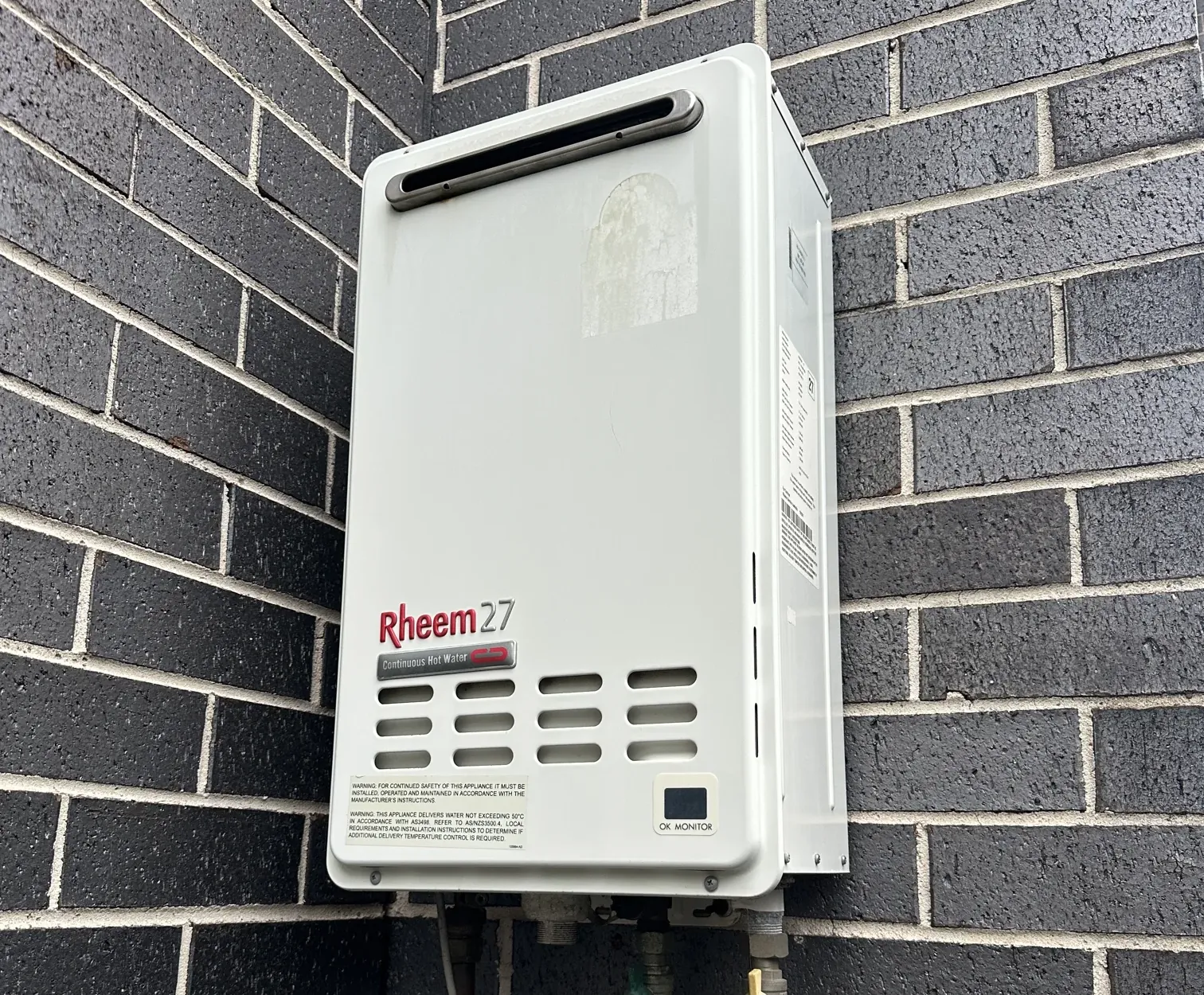 Rheem continuous flow hot water unit with its gas and water connections on a dark brick wall in Canterbury
