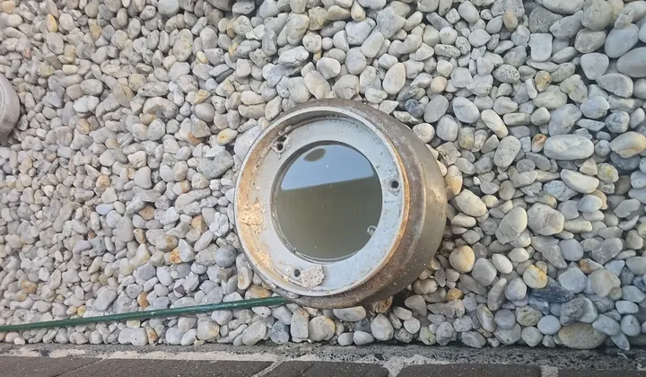 Overflow relief gully set in pebbles with its grate off and water sitting near the top in The Ponds