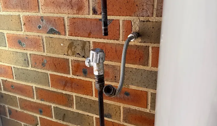 Water pipes, a braided hose and an isolation valve disconnected on a brick wall beside a hot water system in Harrington Park