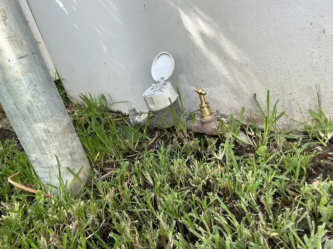 Water meter with its lid open beside a brass tap at the base of a wall in Riverwood