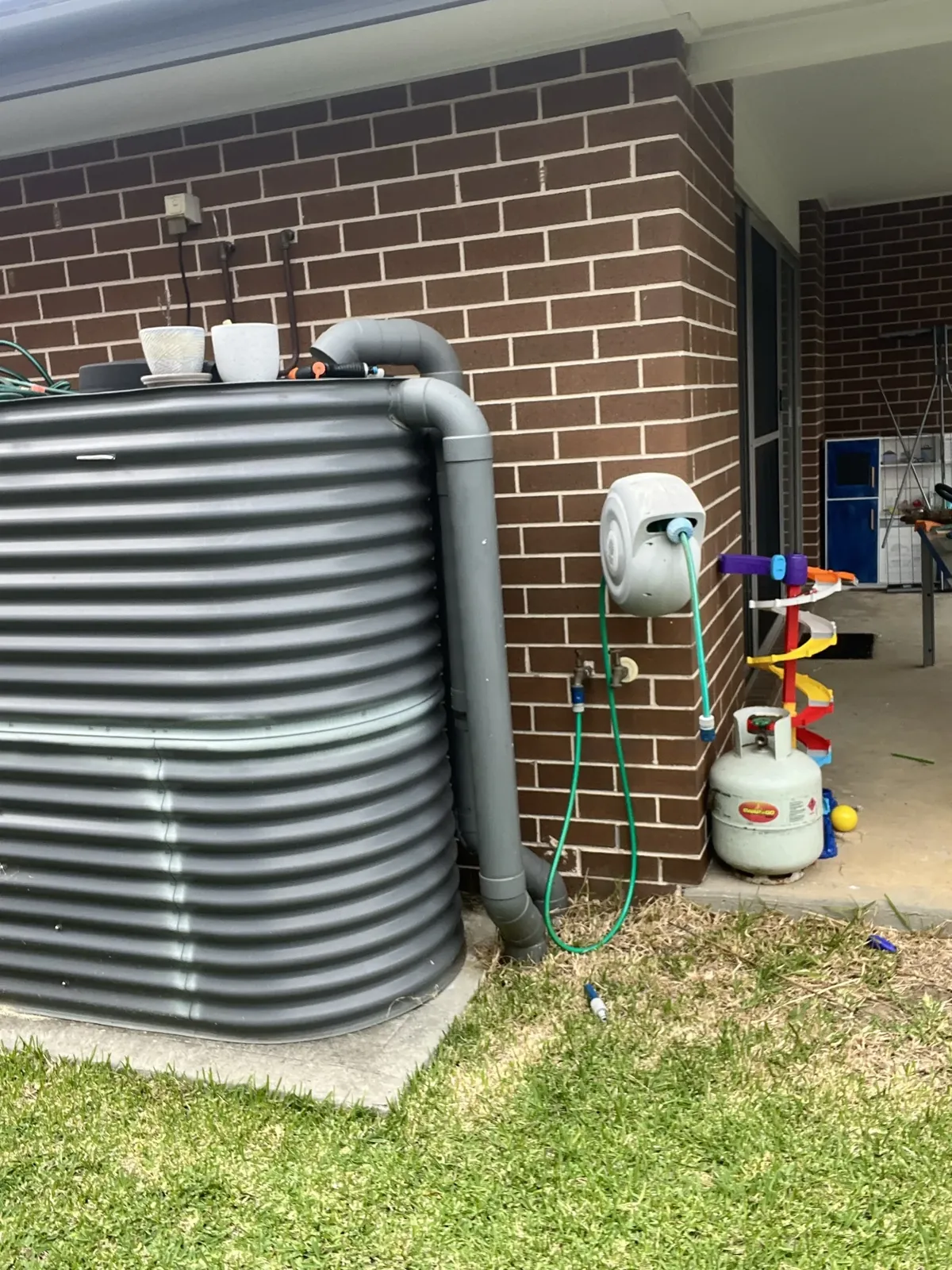 Corrugated rainwater tank with its downpipe and overflow beside a brick house in Gregory Hills