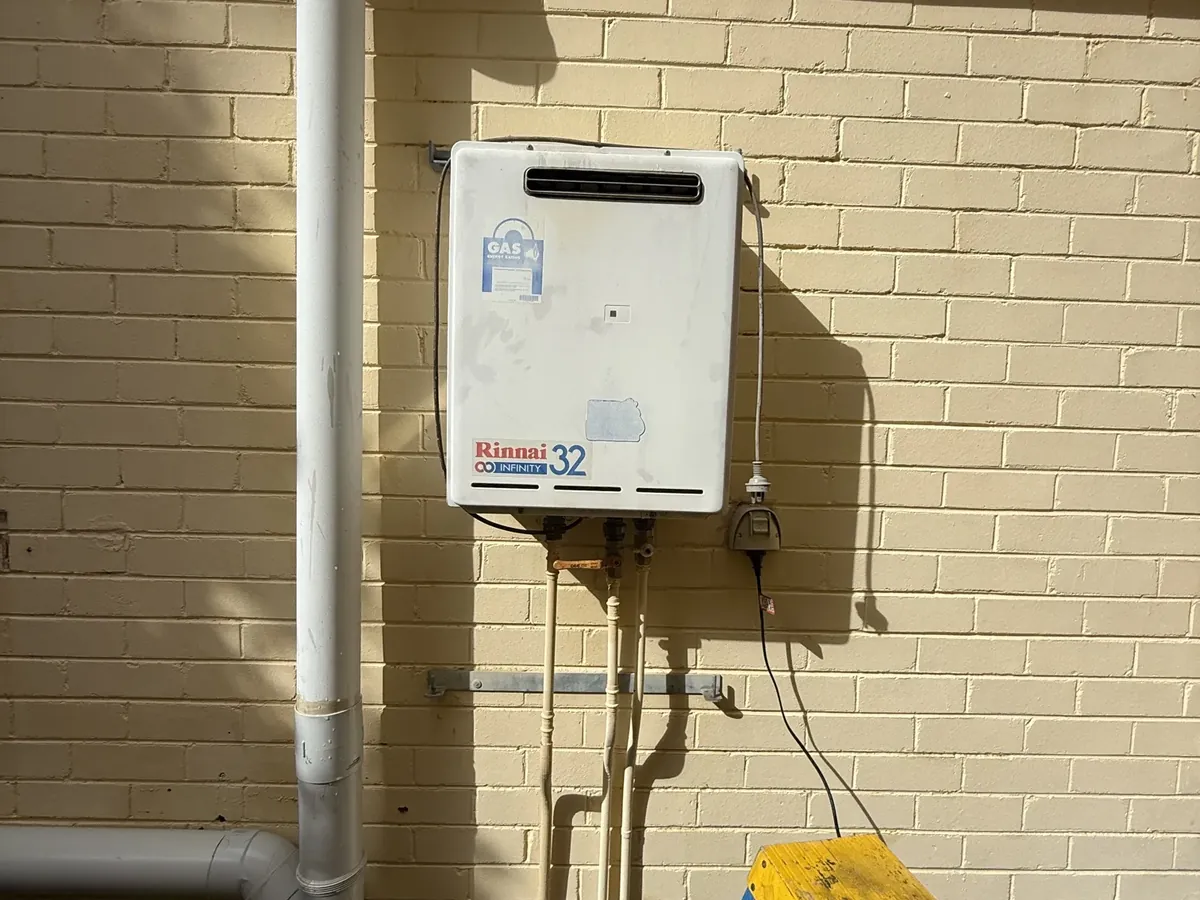Weathered continuous flow gas hot water unit mounted on a cream brick wall between downpipes in Oatley