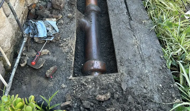 Trench cut along a concrete side path exposing an old clay sewer pipe and junction in Rockdale