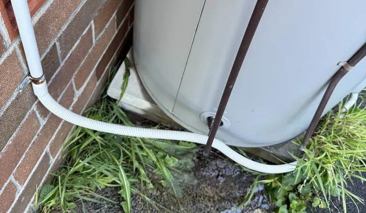 Wet ground and a drain line at the base of a leaking storage tank beside a brick wall in Riverwood