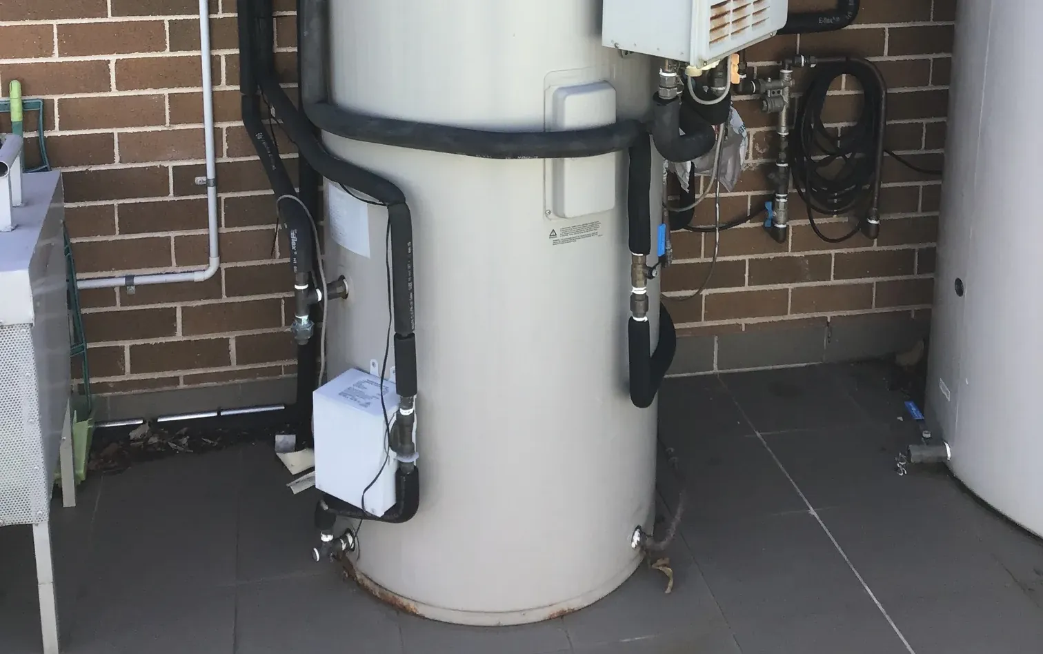 Storage tank with black insulated pipes, valves and a small white box at its base beside a brick wall in Enfield