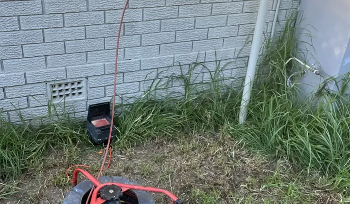 Drain camera reel with its cable fed into a pipe riser beside a brick wall in Dharruk