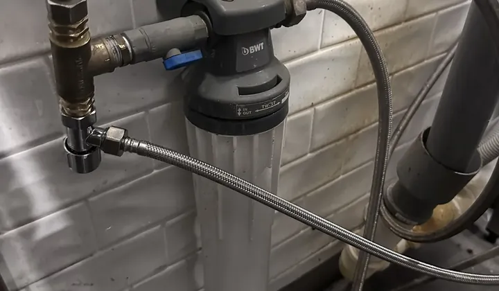 Wall-mounted water filter housing with an isolation valve and braided hoses on a tiled wall beside the drain pipes in a Sydney CBD commercial kitchen