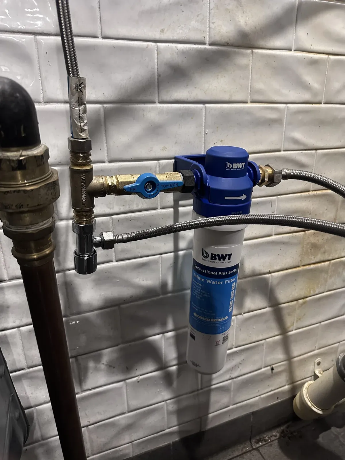 New inline water filter with a fresh cartridge, a brass isolation valve and braided hoses on a tiled wall in a Sydney CBD commercial kitchen
