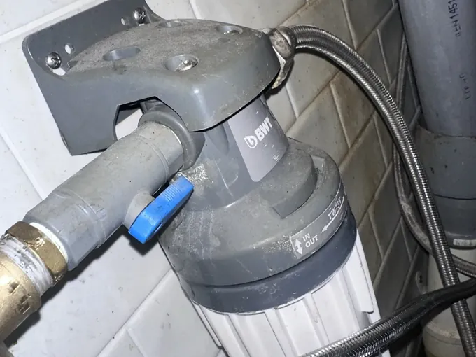 Dusty water filter head with a blue-handled isolation valve and braided hoses on a tiled wall